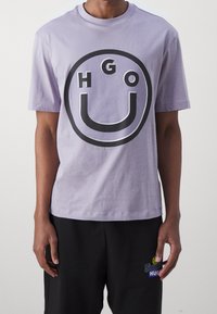 Lilla bomulds t-shirt med et stort sort rundt logo, der viser bogstaverne "HGO" og et smilende "U". Korte ærmer og rund halsudskæring.