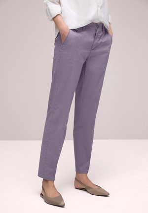 Kobieta w liliowych spodniach o prostych nogawkach z przednimi kieszeniami oraz beżowych, zakrywających piętę balerinach z odkrytymi palcami, ręce w kieszeniach, biała bluzka.