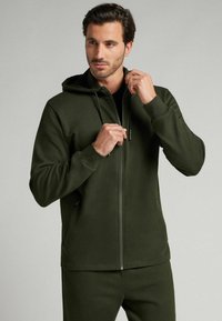 Sudadera con cremallera en verde oliva, con bolsillos frontales, capucha con cordón y puños acanalados; hecha de una suave tela texturada. Combinada con joggers a juego.