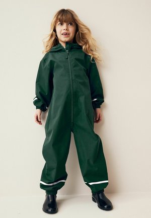 Une combinaison imperméable vert foncé avec une fermeture éclair sur le devant, des poignets ajustables et des rayures blanches au niveau des chevilles. Associée à des bottes en caoutchouc noires et brillantes.