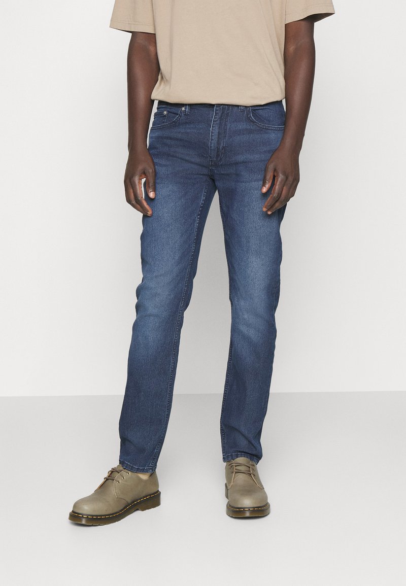 Denim jeans in medium blue with a slim straight fit, featuring five pockets and subtle fading. Paired with light tan lace-up shoes.