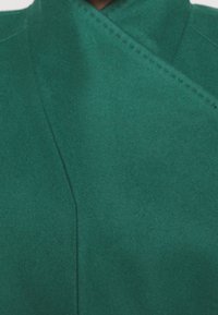 Groene textielstof met een zachte, suède-achtige afwerking. Heeft een wikkelontwerp met zichtbare stiksels langs de naden.