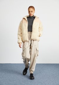 Beige sherpa-jacka med en ljusbrun panel, bärs över en svart- och vitrandig polotröja. Lätta beige cargobyxor och svarta skor.