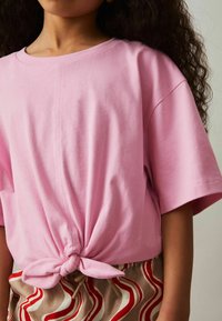 Camiseta de algodón rosa con cuello redondo y mangas cortas, con un dobladillo anudado. Combinada con pantalones cortos estampados en ondas rojas y beige.