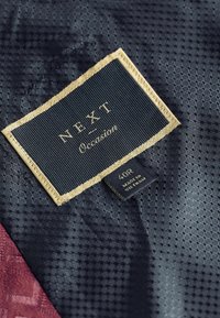 Tela navy con una sutil textura hexagonal, que presenta una etiqueta negra con letras doradas que dice "NEXT Occasion" y detalles de tamaño.