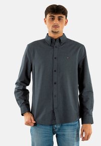 Camisa de manga larga gris oscuro con botones y cuello puntiagudo, con tapeta frontal y un pequeño logo en el pecho. Corte casual y textura suave.