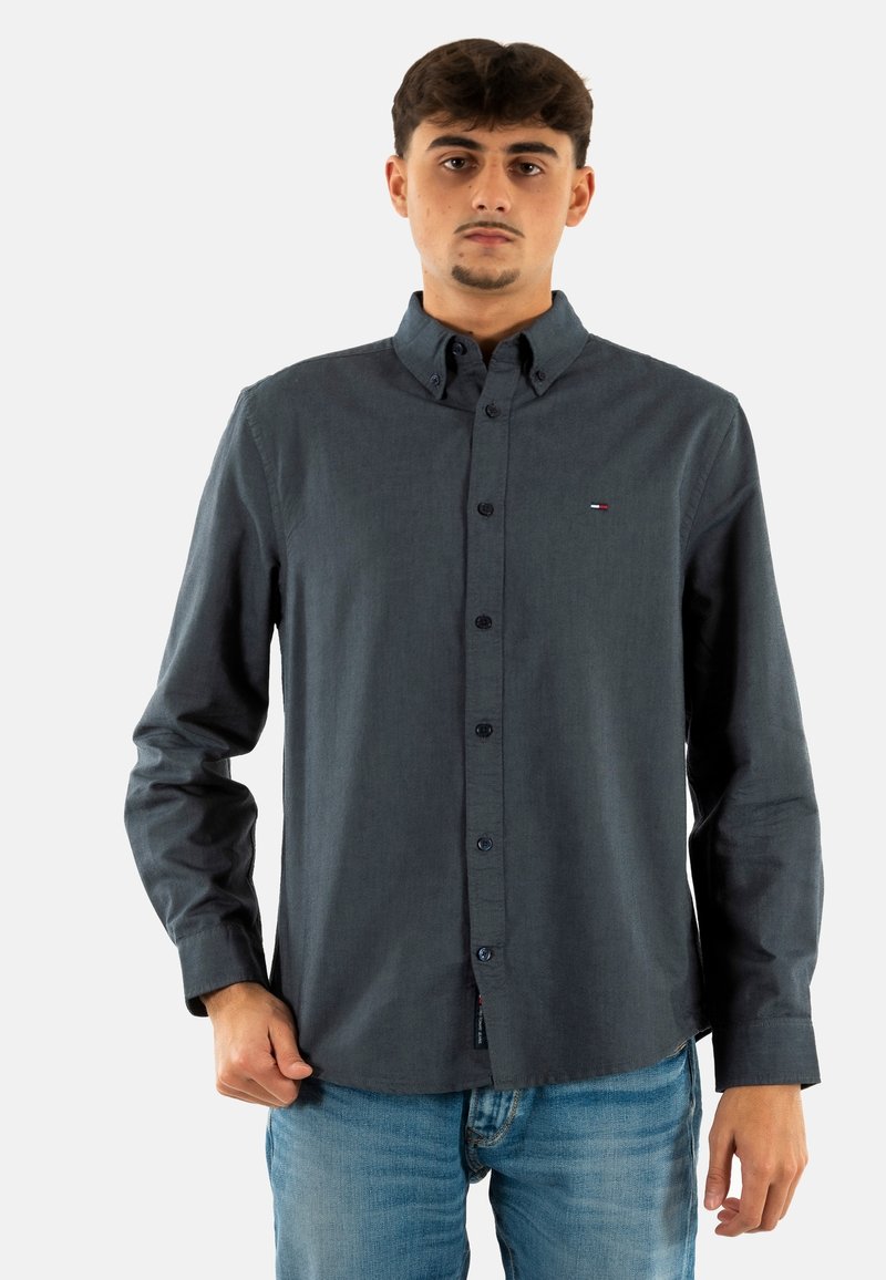 Camisa de manga larga gris oscuro con botones y cuello puntiagudo, con tapeta frontal y un pequeño logo en el pecho. Corte casual y textura suave.