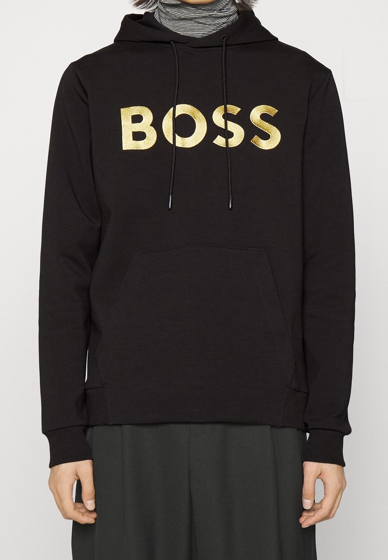 Sweat à capuche noir avec inscription dorée "BOSS" sur la poitrine, porté sur un col roulé rayé, les mains détendues sur les côtés.