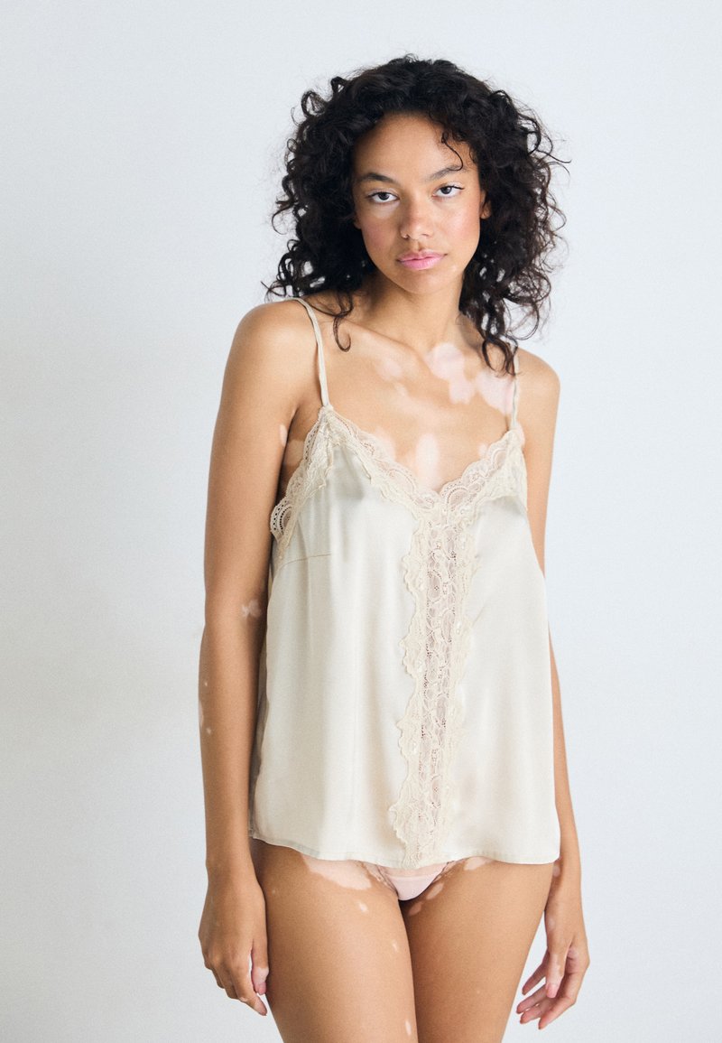 Giovane donna con capelli ricci che indossa un top di pizzo beige e biancheria intima, in piedi contro uno sfondo chiaro e semplice.