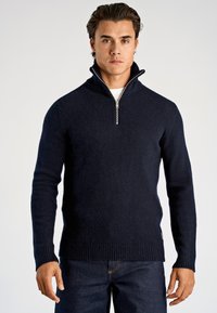 Marineblauer Strickpullover mit Reißverschluss aus strukturiertem Strickmaterial, mit hohem Kragen sowie gerippten Bündchen und Saum. Über einem weißen Hemd getragen.