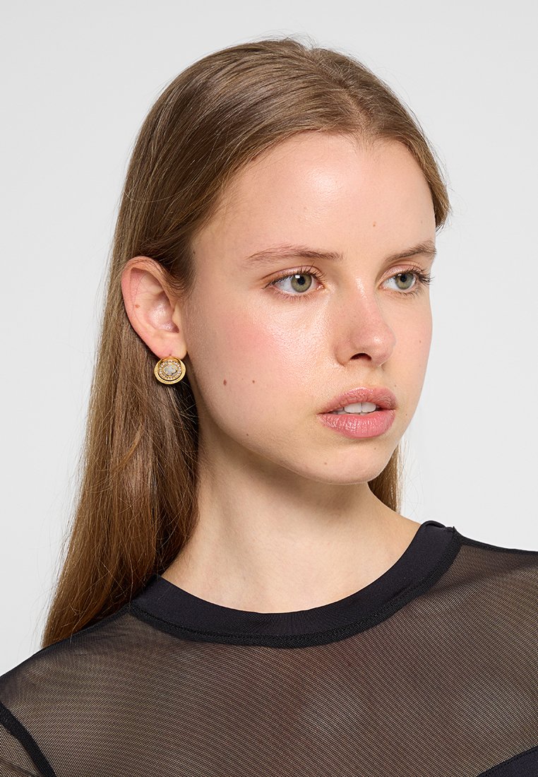 Boucles d'oreilles studs en ton doré avec un design circulaire et une surface texturée, ornées d'une pierre centrale. Portées par un modèle.
