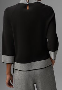 Svart topp med hög krage, som har en nyckelhålsknäppning bak. Utsmyckad med en grå randig kant vid ärmar och nederkant.