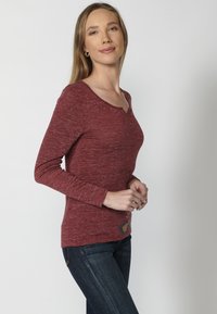 Bordó hosszú ujjú felső, V-nyakkal, amely diszkrét fémszínekkel van díszítve a nyakrésznél, és egy dekoratív folttal a bal alsó részen.
