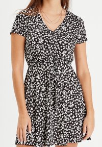 Femme portant une robe noire courte avec un imprimé floral blanc, des manches courtes, un devant boutonné, une ceinture à la taille et des colliers en or superposés.