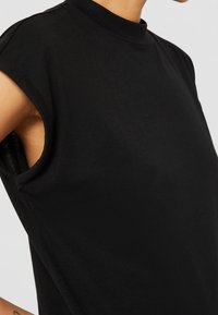 Buste supérieur portant un haut noir sans manches avec de larges emmanchures et un col montant sur un fond uni.