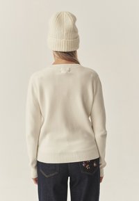 Pulover de culoare crem deschis, cu manșete și tiv cu nervuri, purtat cu blugi de denim închis, care au broderie florală pe buzunarele din spate. Căciulă tricotată.