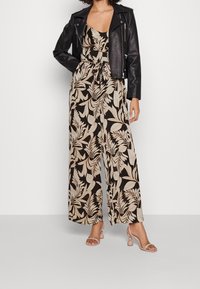 Mono negro con estampado de hojas, con cuello con volantes y cintura ajustada, combinado con una chaqueta de cuero negra ajustada y sandalias de tacón nude.