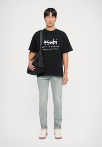 Must kollane oversize T-särk valge brändi logoga, helesinised kulunud teksad ja beežid spordijalatsid, hoides musta rihmaga kotti.