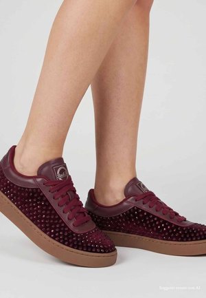 Gambe con sneakers rosso scuro con dettagli borchiati e suole in gomma, in piedi su uno sfondo grigio chiaro uniforme.