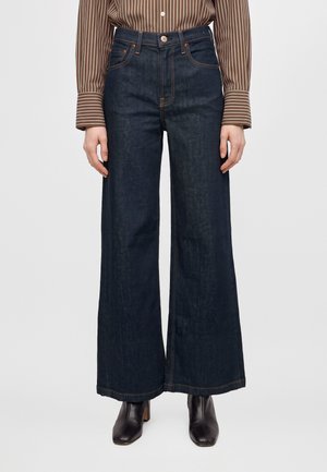 Mannequin portant un jean indigo foncé taille haute à jambes larges, une chemise marron à rayures et des bottines noires.