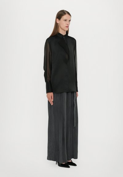 Black sheer long-sleeve blouse with a collar, paired with high-waisted gray wide-leg trousers. Model wears black high heels.