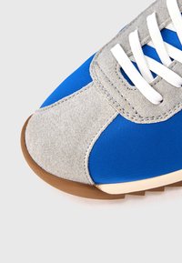 Detailný záber modrej tenisky so sivými semišovými prekladmi, bielymi šnúrkami a hnedou gumovou podrážkou na bielom pozadí.