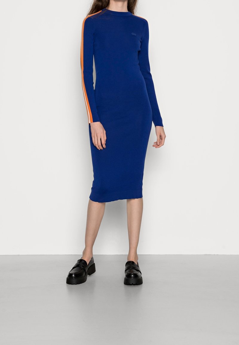 Tajtat blå stickad klänning med långa ärmar, prydd med orange sidostreck. Knälång med rund halsringning. Buren med svarta platåskor.