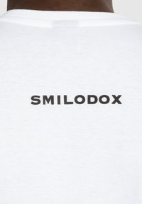 Hvid bomulds t-shirt med et sort trykt logo "SMILODOX" på ryggen, med en klassisk rund halslinning og glat tekstur.