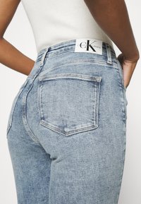 Jeans de mezclilla de un azul claro con una cintura alta, diseño de cinco bolsillos y una etiqueta blanca "Calvin Klein Jeans" en la parte posterior. Textura suave.