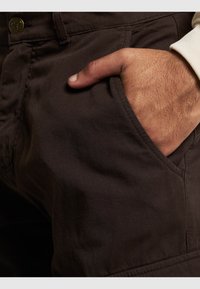 Donkerbruine stoffen broek met een voorzak, gekenmerkt door een gladde textuur en verstevigde naden, met een zichtbare knoopdetail.