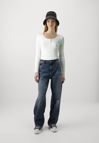 Bluză albă cu mâneci lungi și detalii cu butoane, asociată cu blugi de tip wide-leg albastri, distruși, și o pălărie de tip bucket neagră. Adidașii au completat ținuta.