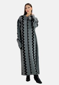 Lang grijze sweater jurk met zwarte zigzagpatronen in het midden, voorzien van brede mouwen en een geribde halslijn. Gekleed met zwarte laarzen.