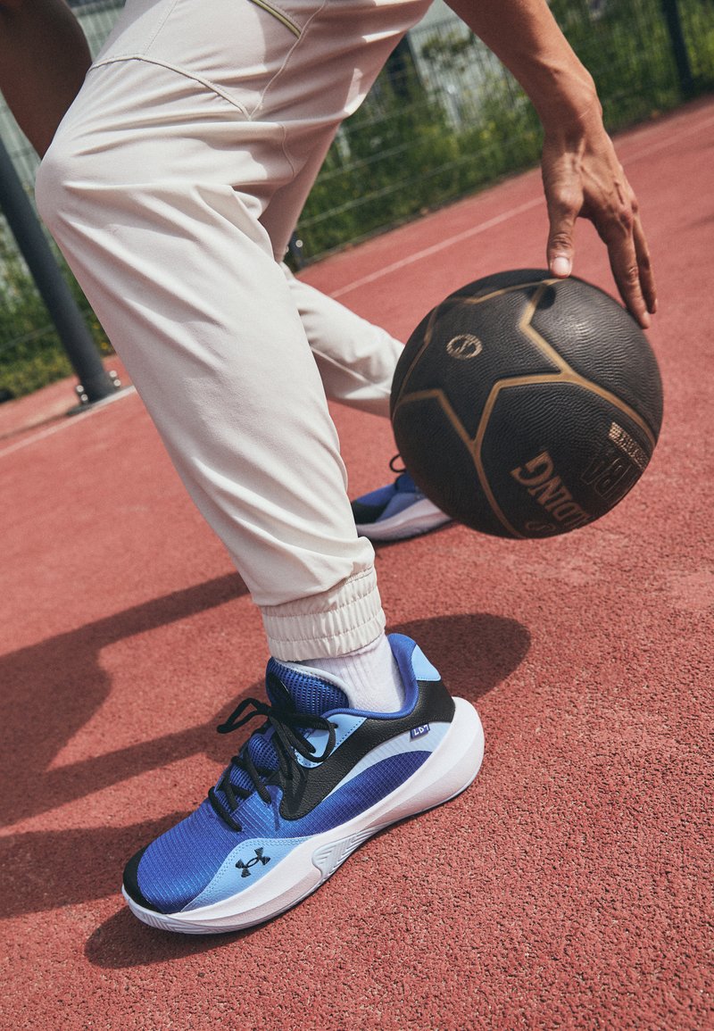 Chaussures de sport bleues avec un swoosh noir, tige texturée et semelle blanche. Pantalons blancs avec des poignets élastiques, drapés sur un sol de court rouge.