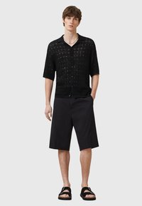 Camisa negra de manga corta y punto abierto con cuello, botones en el frente y diseño texturizado, combinada con pantalones cortos negros hasta la rodilla y sandalias.