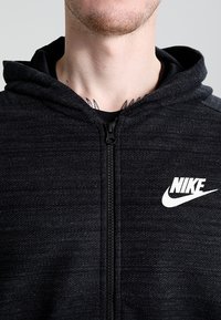 Svart zip-up hoodie i texturerat tyg, med en vit Nike-logga på bröstet och en huva med dragsko.