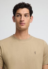 Beżowa bawełniana koszulka z okrągłym dekoltem, krótkimi rękawami i małym haftowanym logo na lewej piersi. Gładka tekstura.