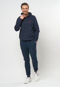 Marineblauwe hoodie met trekkoord, kangoeroezak en klein logo. Gecombineerd met bijpassende joggers met elastische boorden en zakken. Witte sneakers.
