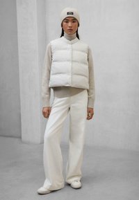 Chaleco de plumas blanco, suéter de punto beige suave, pantalones blancos de pierna ancha. La modelo lleva un gorro de punto ligero; diseño minimalista con tonos neutros.