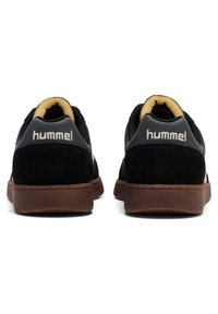 Baskets noires avec une tige en suède et mesh, semelle en caoutchouc marron, et un subtil logo Hummel sur le talon. Design minimaliste avec une forme arrondie.