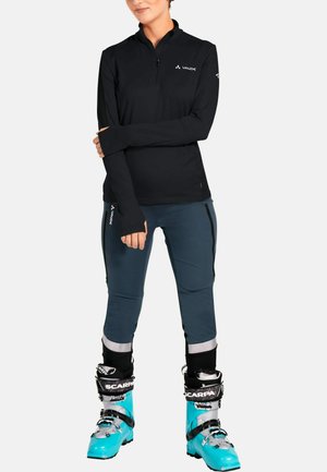Zwarte lange mouwen zip-up top met duim gaten, blauwe softshell broek met zijkanten ritsen, gecombineerd met turquoise skibotten.