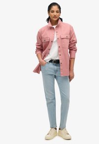 Veste rose à boutons avec deux poches poitrine, t-shirt blanc, jeans bleu clair et baskets beiges. Texture lisse et coupe classique.