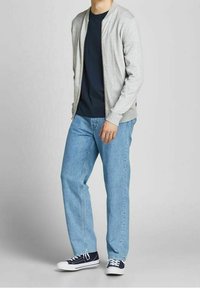 Man som bär en ljusgrå zip-up jacka, svart t-shirt, ljusblå jeans och svarta sneakers och står mot en enfärgad bakgrund.