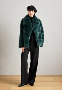 Türkisfarbener Kunstpelzmantel mit breitem Kragen, schwarzem Oberteil und schwarzen Hosen. Das Modell trägt eine schwarze Baskenmütze und präsentiert ein strukturiertes, geschichtetes Design.