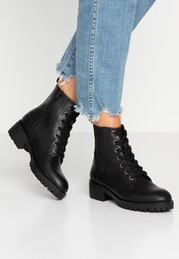 Bottines en cuir noir à lacets, avec œillets en métal contrastants, semelle épaisse et talon légèrement rehaussé. Un pantalon en denim usé complète le look.
