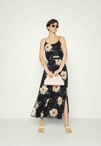 Vestido maxi preto com alças finas, apresentando grandes flores em pêssego e creme, fenda lateral, combinado com uma clutch bege e sandálias.