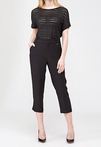 Černý průhledný pruhovaný top s širokými rukávy v kombinaci s černými kalhotami s vysokým pasem a zkrácenou délkou. Modelka má na sobě černé vysoké podpatky, na jednobarevném pozadí.