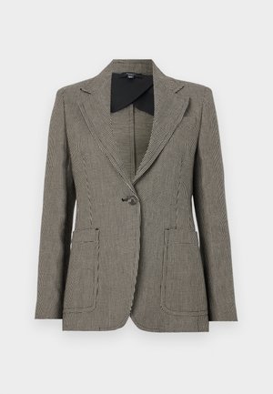 Henkilö yllään ruudullinen bleiseri, musta toppi, valkoiset leveälahkeiset housut, mustat ballerinat ja musta käsilaukku kädessä.