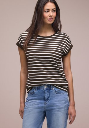 Kobieta o długich brązowych włosach, ubrana w brązowo-biały paskowany t-shirt z krótkim rękawem i jasnoniebieskie dżinsy, stojąca na jednolitym tle.