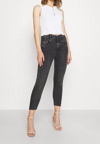 Vaqueros negros ajustados de cintura alta, con bolsillos delanteros y detalles de desgaste sutil, combinados con una blusa blanca corta con bordes ondulados.