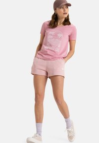 Camiseta rosa de manga corta con gráfico de palmera y texto "Roxy Desde 1990", combinada con pantalones cortos rosa claro. Modelo lleva una gorra y zapatillas.